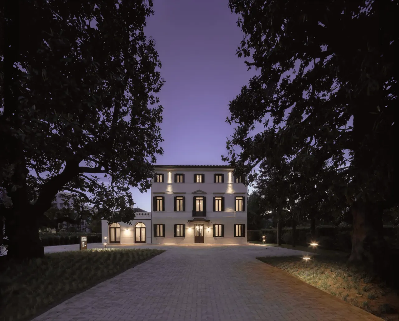 Historic villa facade lit at dusk at the end of a tree-lined driveway