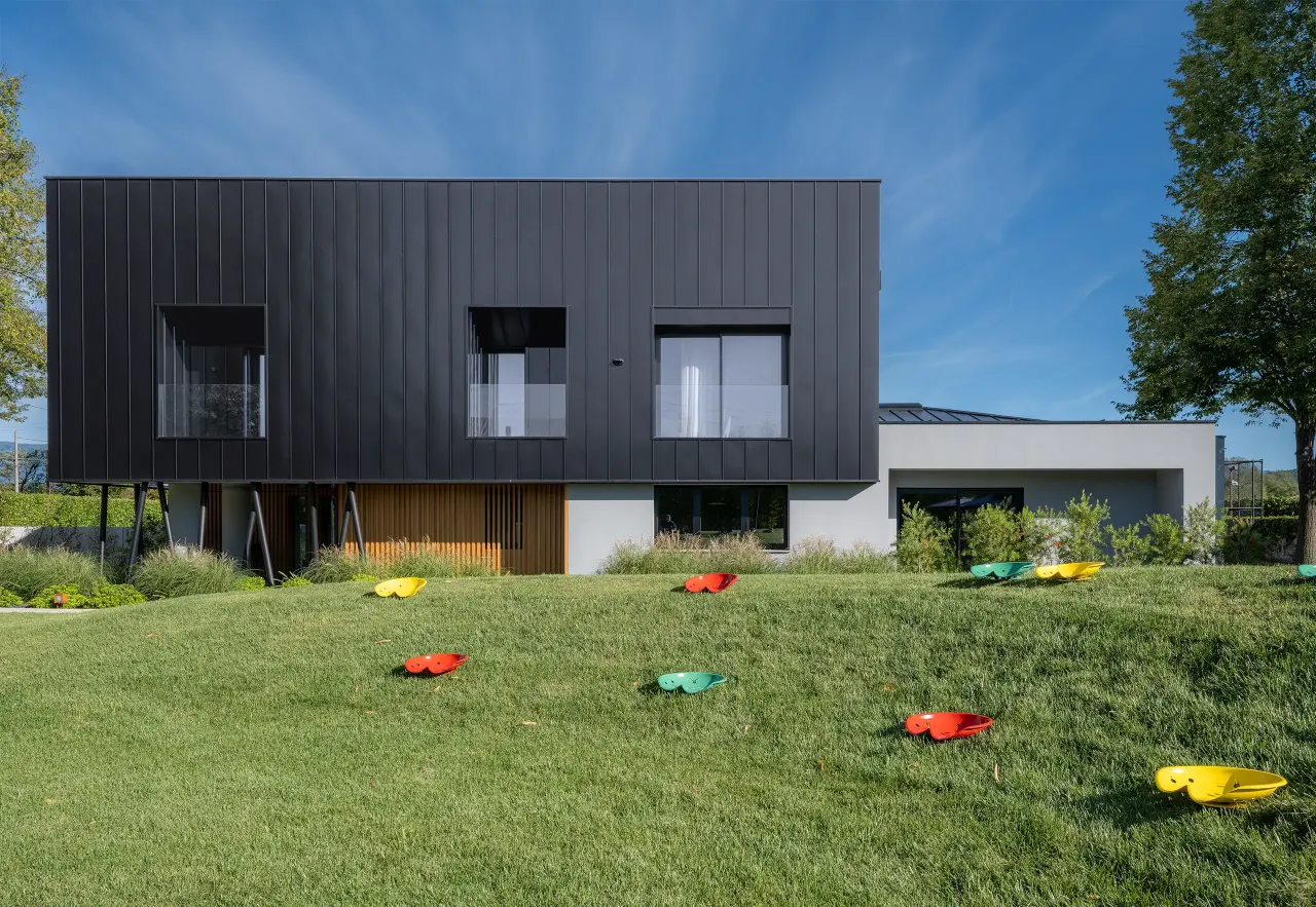 Dark metal-clad building with white ground floor above a lawn with colourful seats