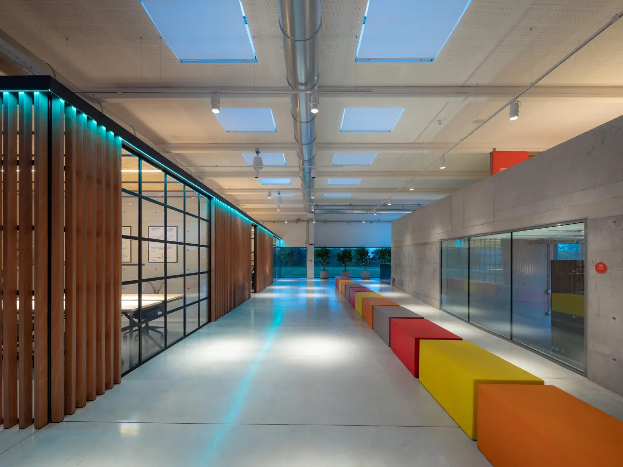 Long corridor with coloured benches, concrete walls and glazed offices