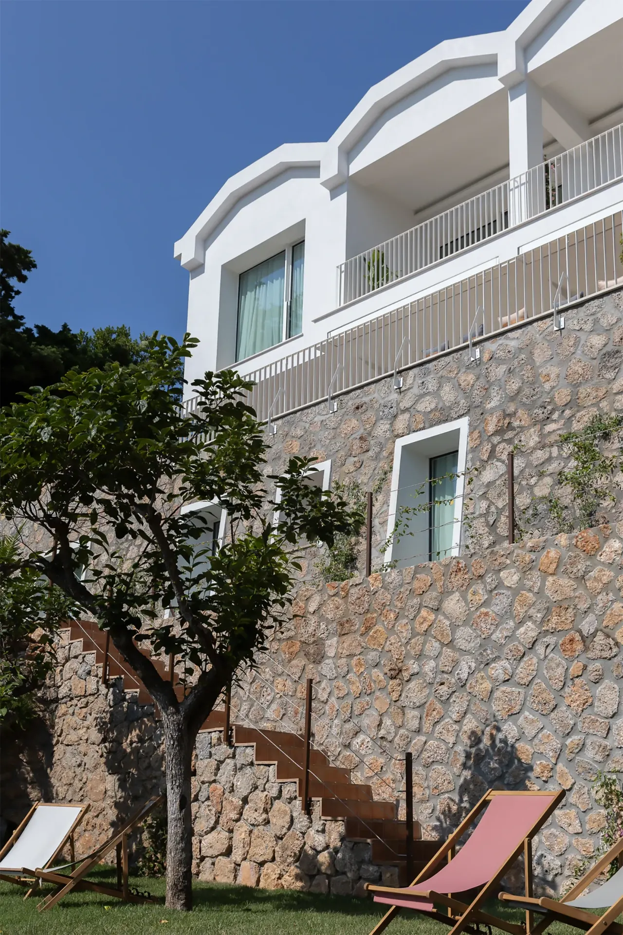 White villa above a stone retaining wall with deck chairs on the lawn